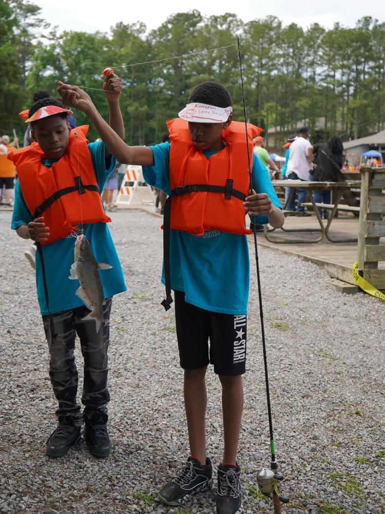 Two boys stand near a lake and wear lifejackets. They hold up a fish on fishing line.