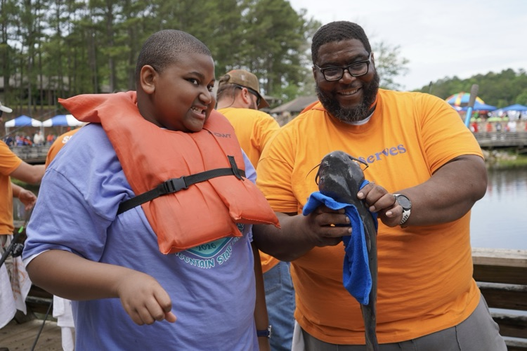 A boy and a man stand together outside. The man holds a catfish in his hands.