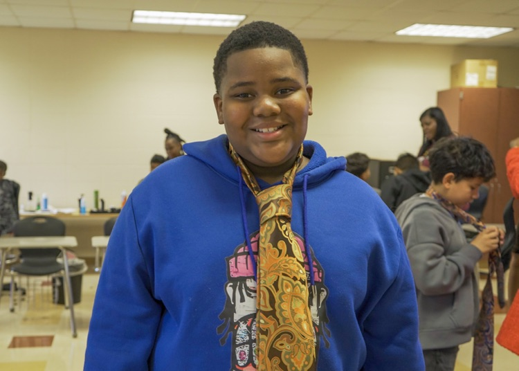 A boy stands in a room and smiles. He wears a tie over his clothes 