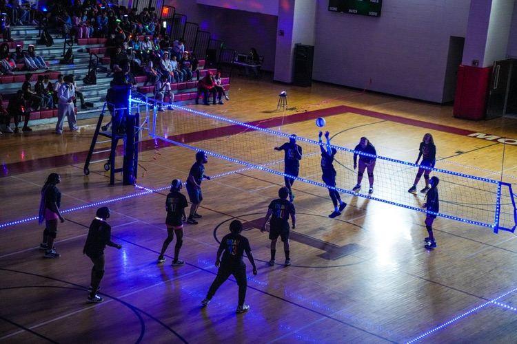 Students play volleyball inside a dark gym. The gym is decorated with colorful lights.