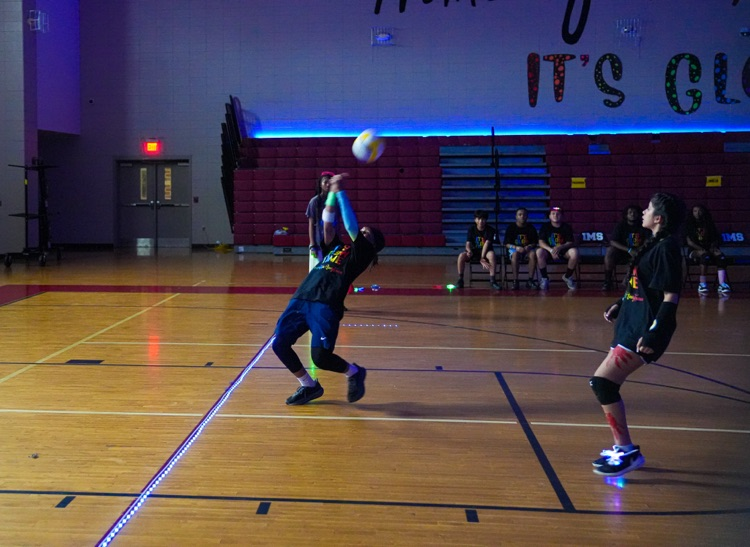 A student hits a volleyball over his head while playing in a dark gym decorated with glowing lights.