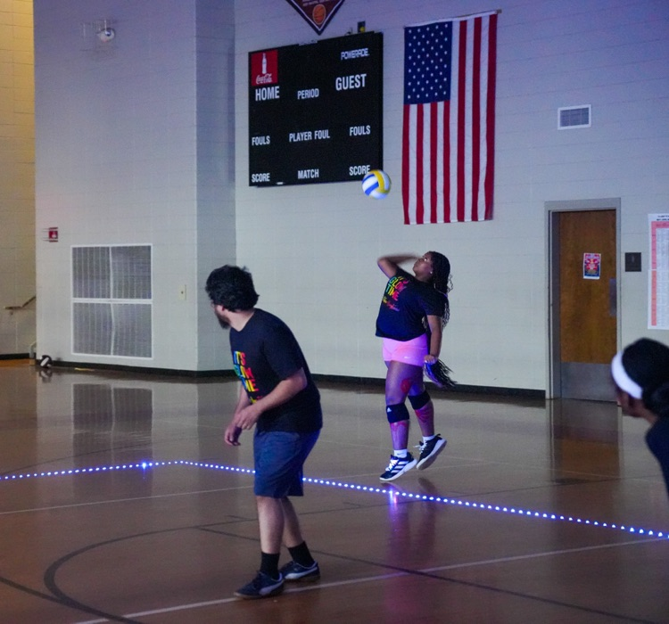 A student serving a volleyball while playing in a dark gym decorated with glowing lights.