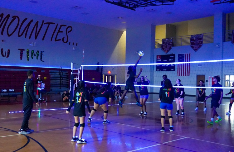 A student hits a volleyball over a net while playing in a dark gym decorated with glowing lights.