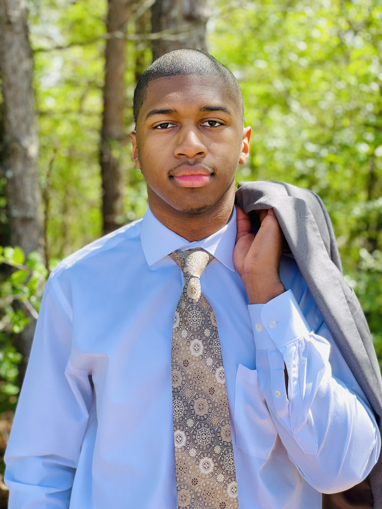 Shades Valley High School senior Abraham Clark smiles for a photo outdoors. He wears a dress shirt and tie and holds a suit jacket in his hand.