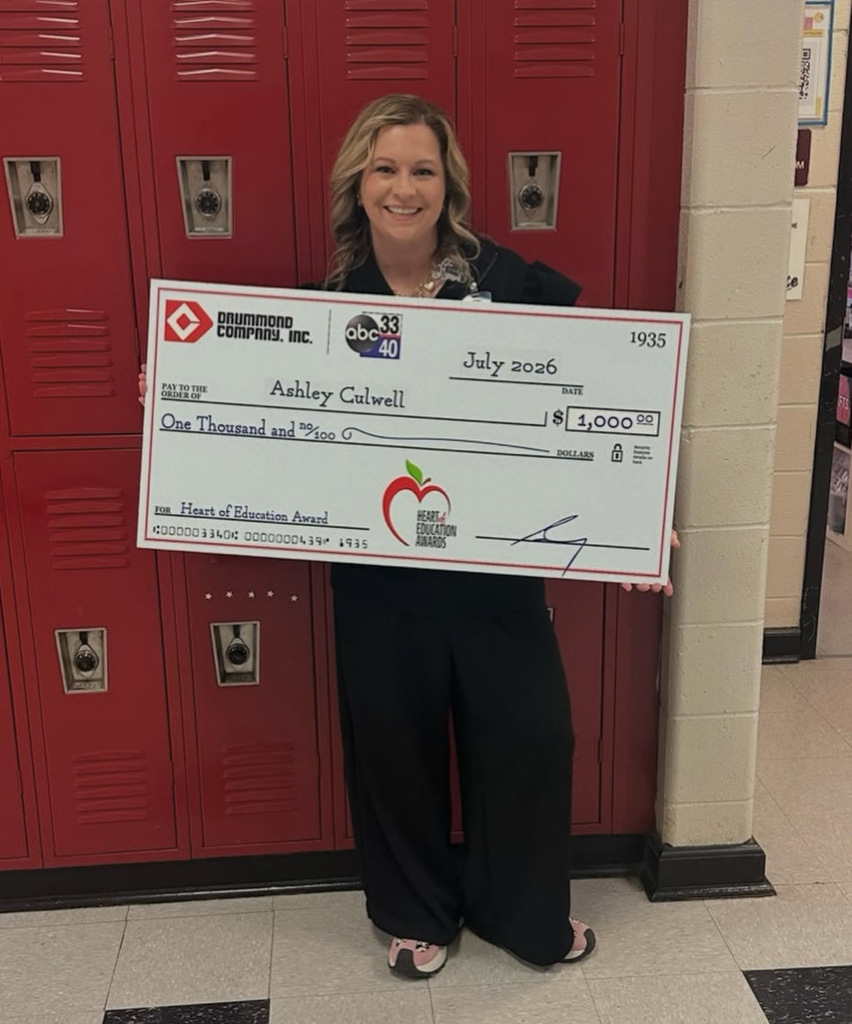 Mrs. Culwell, visual arts teacher at Shades Valley High School smiles for a photo in the school hallway. She holds a large check for $1,000.