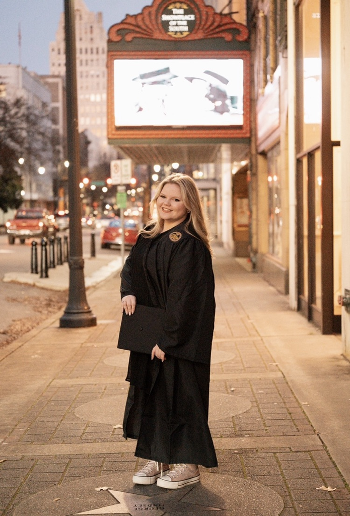 A girl smiles while standing on a sidewalk. She wears a cap and gown.