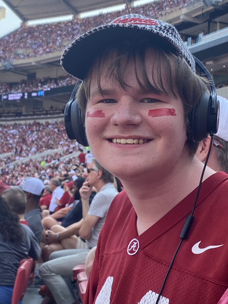 Joseph Hamrick smiles for a photo while sitting in a crowded football stadium. He wears an Alabama football jersey.