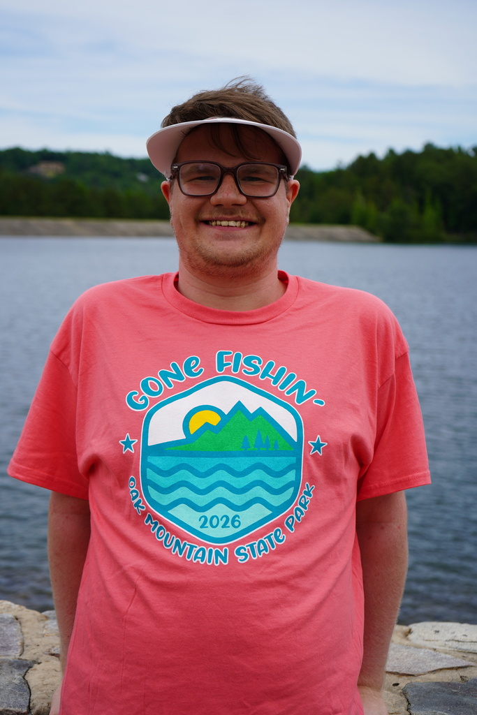 Joseph Hamrick smiles for a photo. He wears a bright pink shirt that says "Gone Fishin' Oak Mountain State Park" and stands in front of a lake.