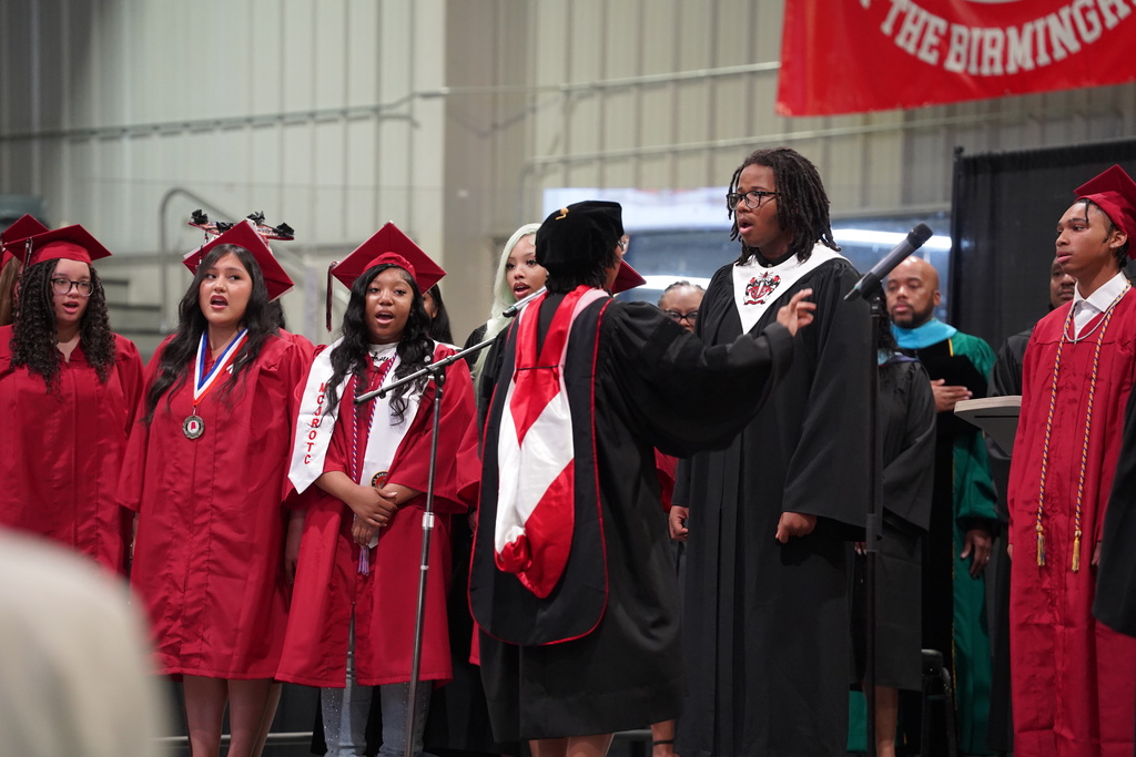 Students wear gowns and sing on stage.