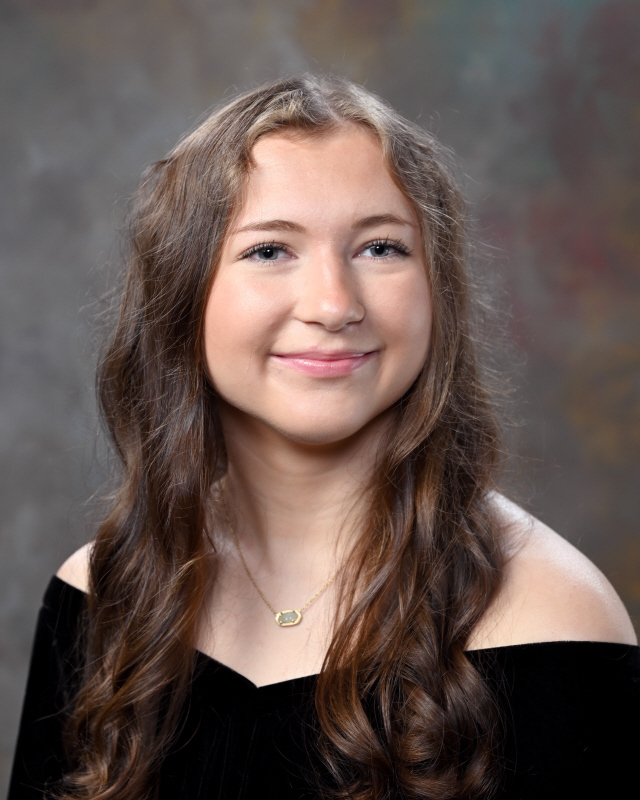 Oak Grove student Allie Rhine wears a black shirt and smiles while standing in front of a gray background.