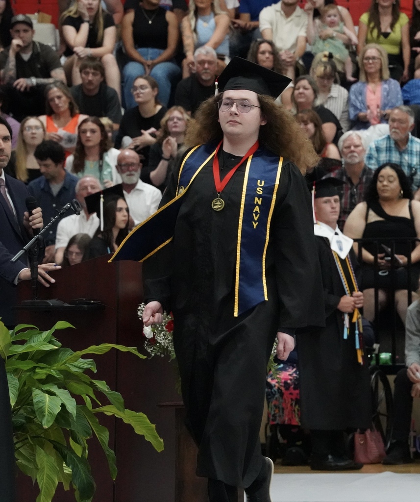 A student walks in front of a gym filled with people during Oak Grove's graduation ceremony. He wears a cap and gown. 