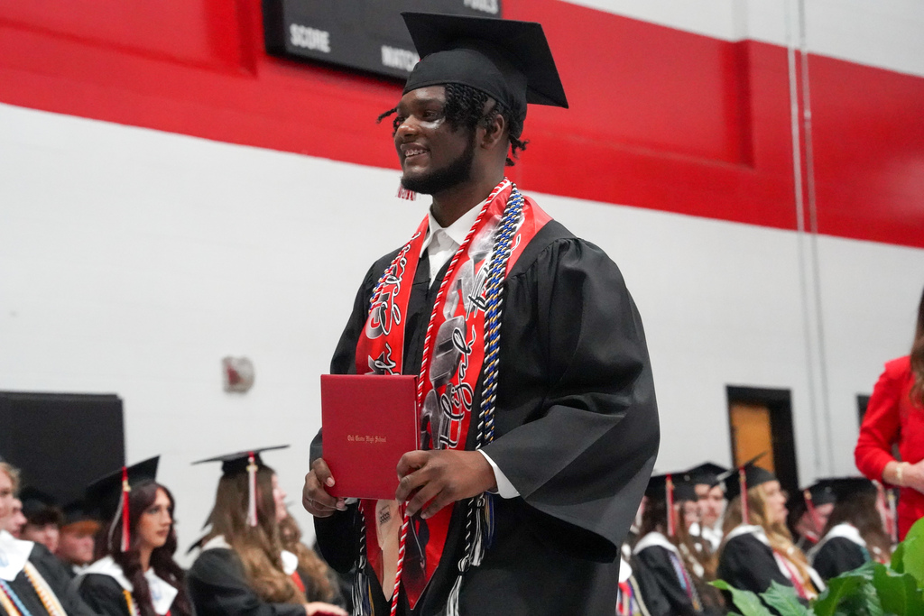 A student smiles while holding a diploma and wearing a cap and gown during Oak Grove High School's graduation ceremony.