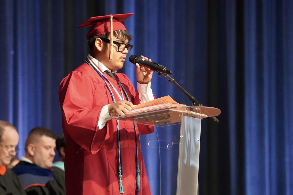 A student speaks into a microphone during the Pinson Valley High School graduation ceremony. He wears a red cap and gown.