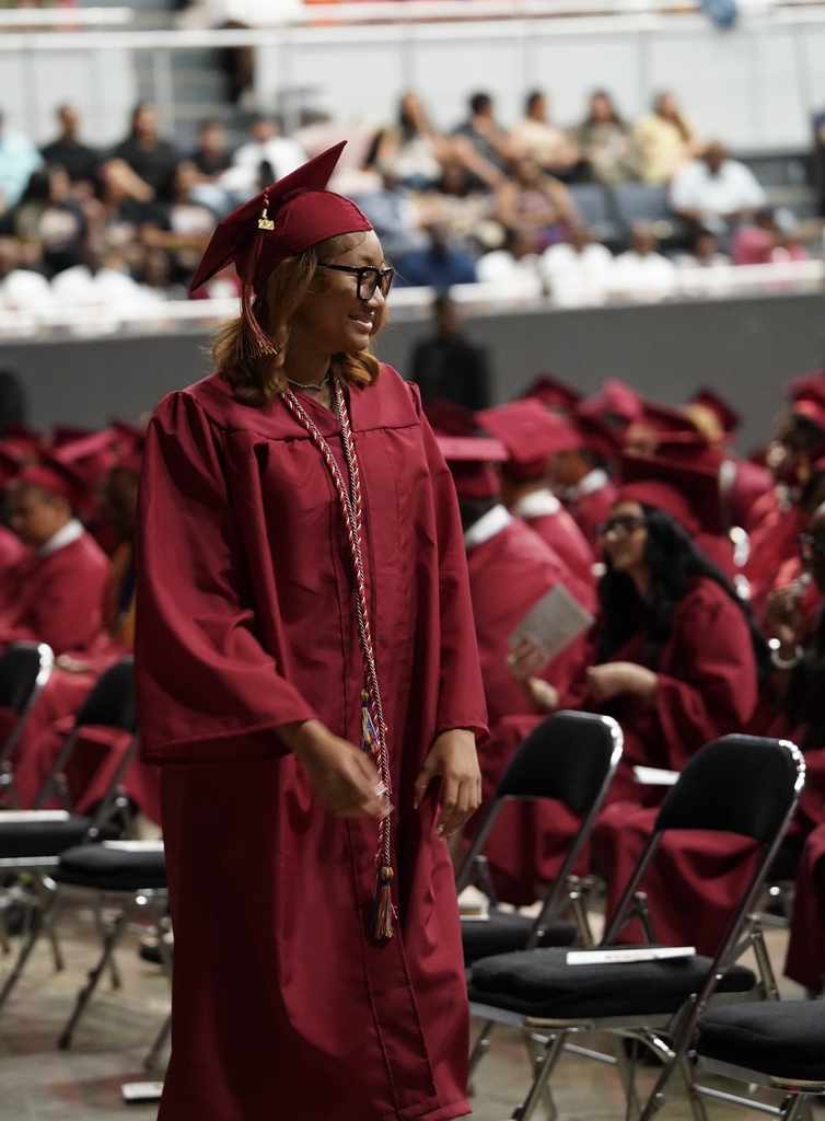 A girl wearing a red cap and gown smiles while walking in an auditorium during her high school graduation ceremony.