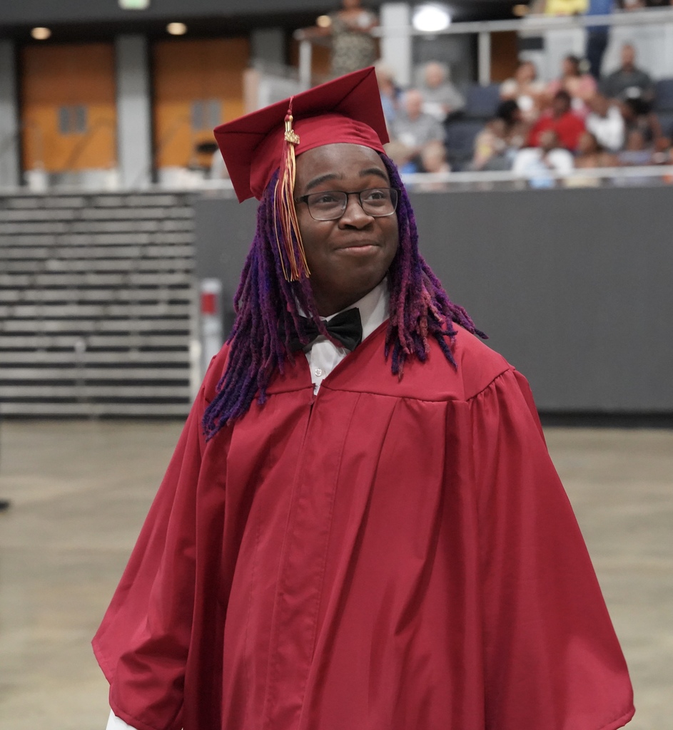 A student wearing a red cap and gown smiles while entering an auditorium for his high school graduation ceremony.
