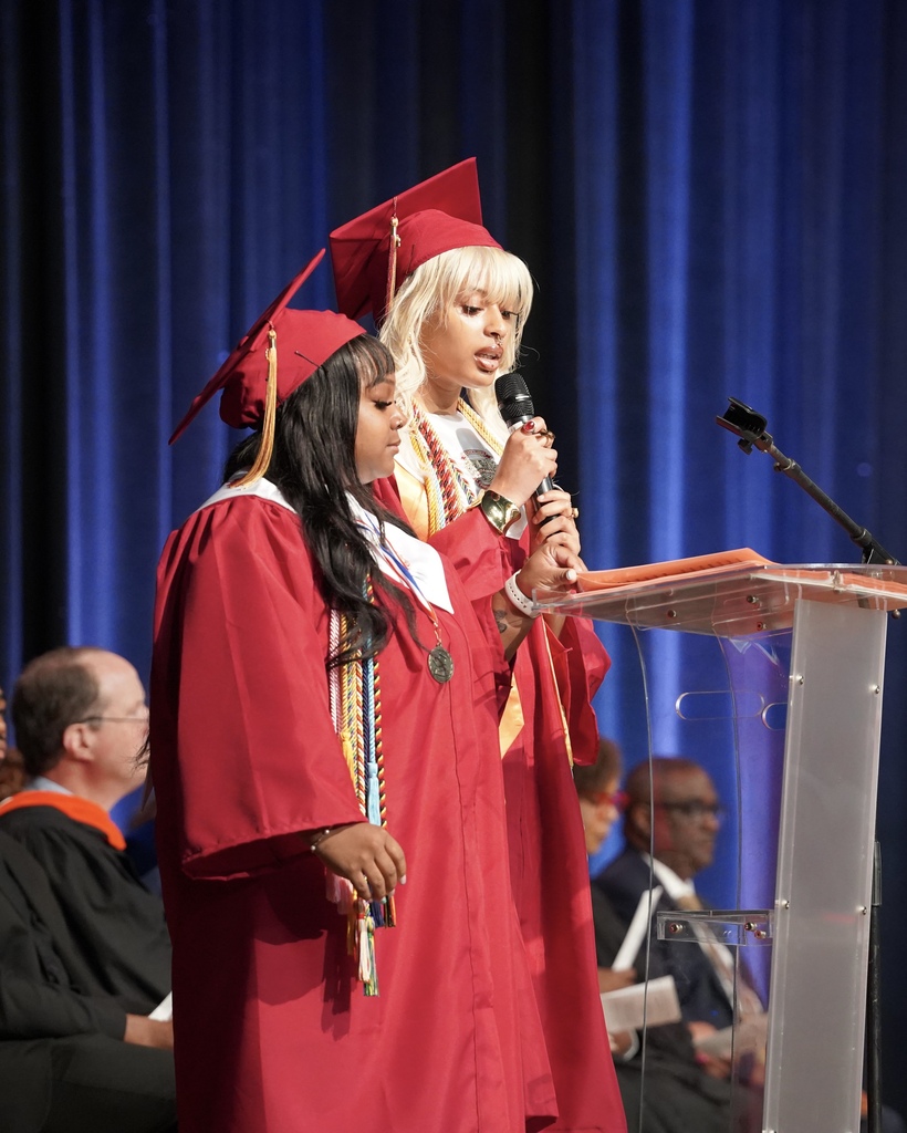 Two students stand at a podium during their high school graduation ceremony. They both wear red caps and gowns. One of the students holds a microphone and speaks into it.