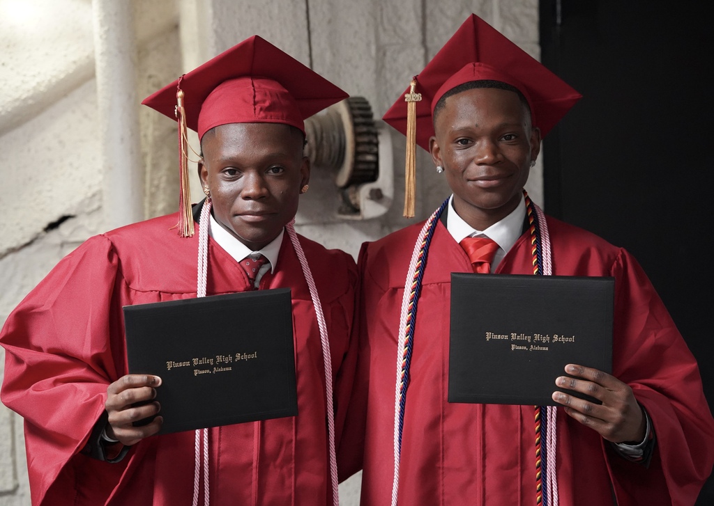 Twin brothers wearing red caps and gowns smile for a photo. They hold their high school diplomas in their hands.