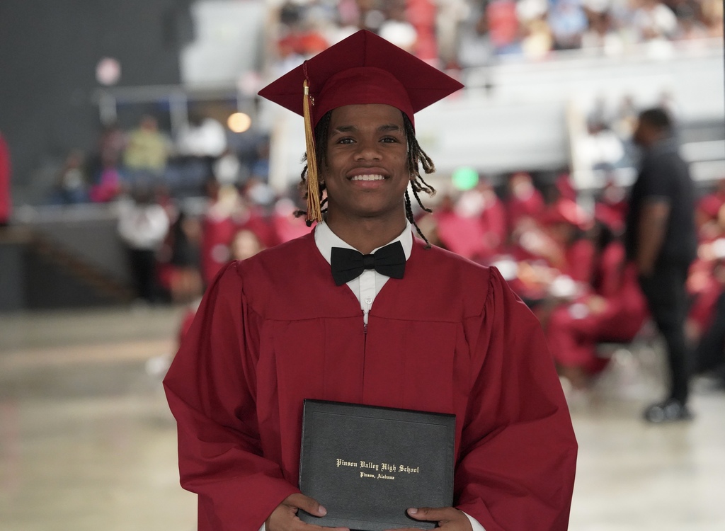 A graduate wearing a red cap and gown smiles and holds his high school diploma.