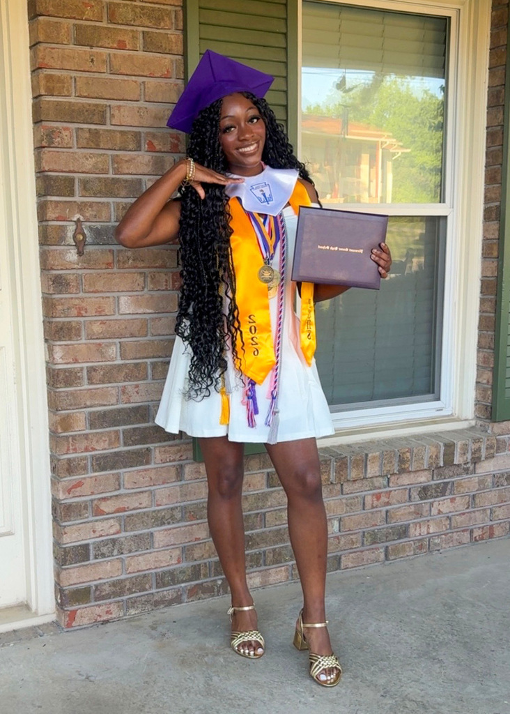 Pleasant Grove High School graduate Laura Norris smiles for a photo. She stands outside a home wearing a white dress and a purple graduation cap and graduation cords.