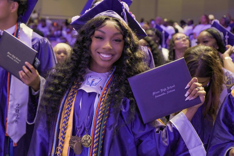 A Minor High School graduate smiles and holds up her diploma during the graduation ceremony. She wears a purple cap and gown
