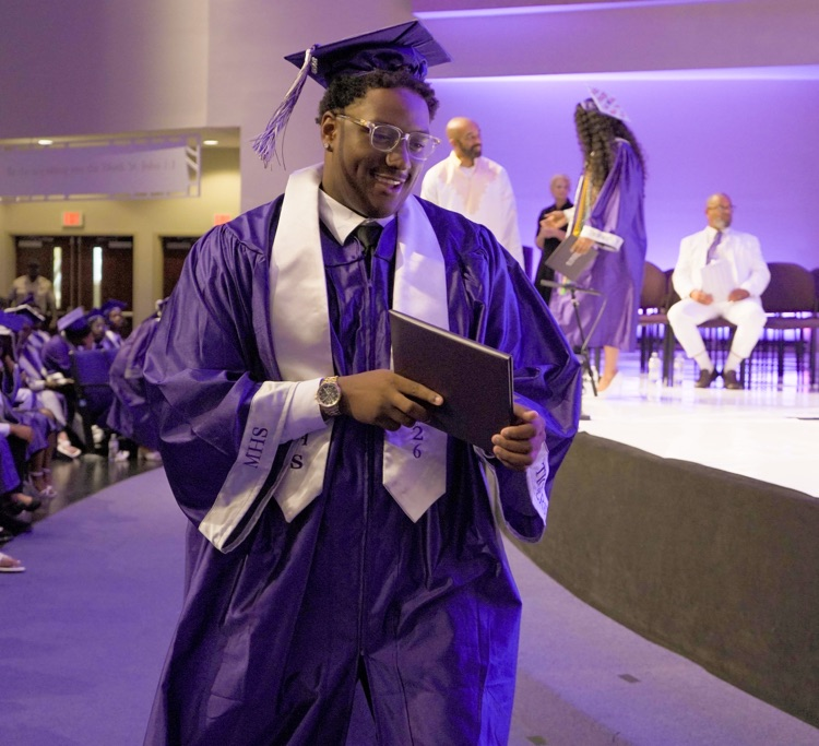A Minor High School graduate smiles as he leaves the stage with his diploma. He wears a purple cap and gown