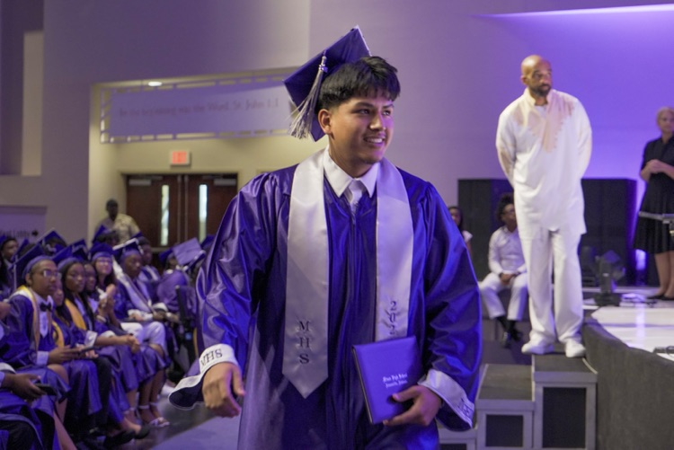 A Minor High School graduate smiles after receiving his diploma. He wears a purple cap and gown