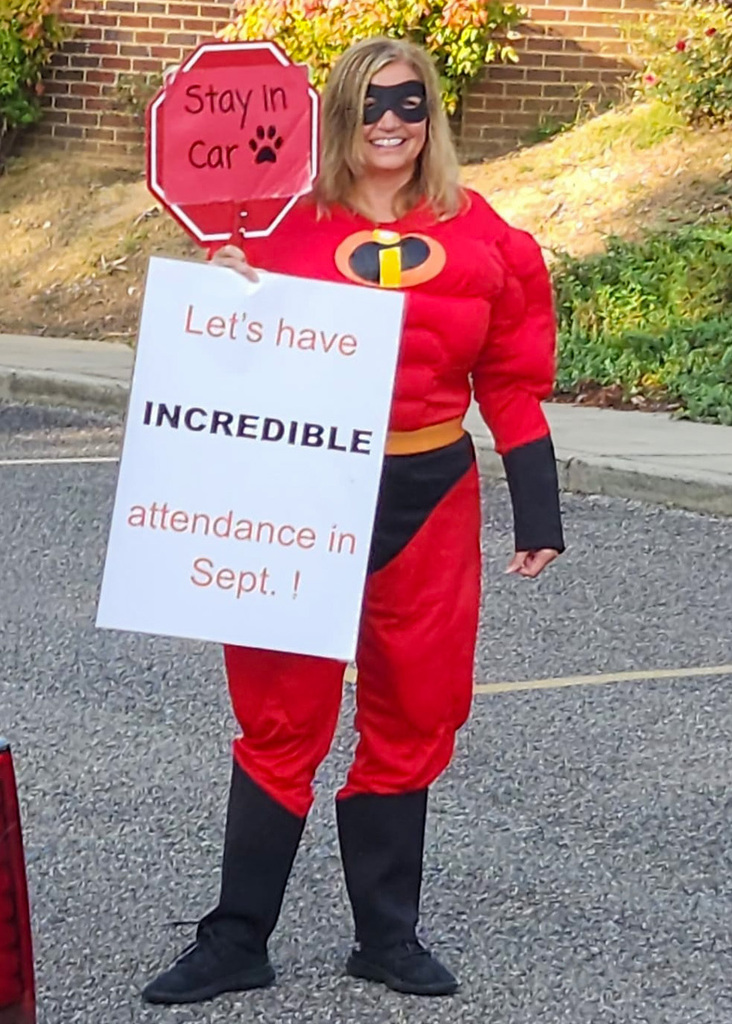 Snow Rogers principal Kristi Long wears an Incredibles Super Hero costume and stands outside the school. She holds a stop sign that says "Stay in Car" and a sign that says "Let's have incredible attendance in Sept."