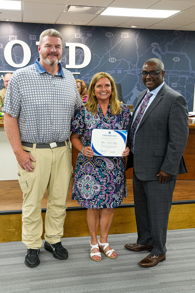 Superintendent Dr. Walter Gonsoulin, Kristi Long and Kristi's husband stand together inside the JEFCOED board room and smile for a photo. Kristi holds a certificate celebrating her retirement.