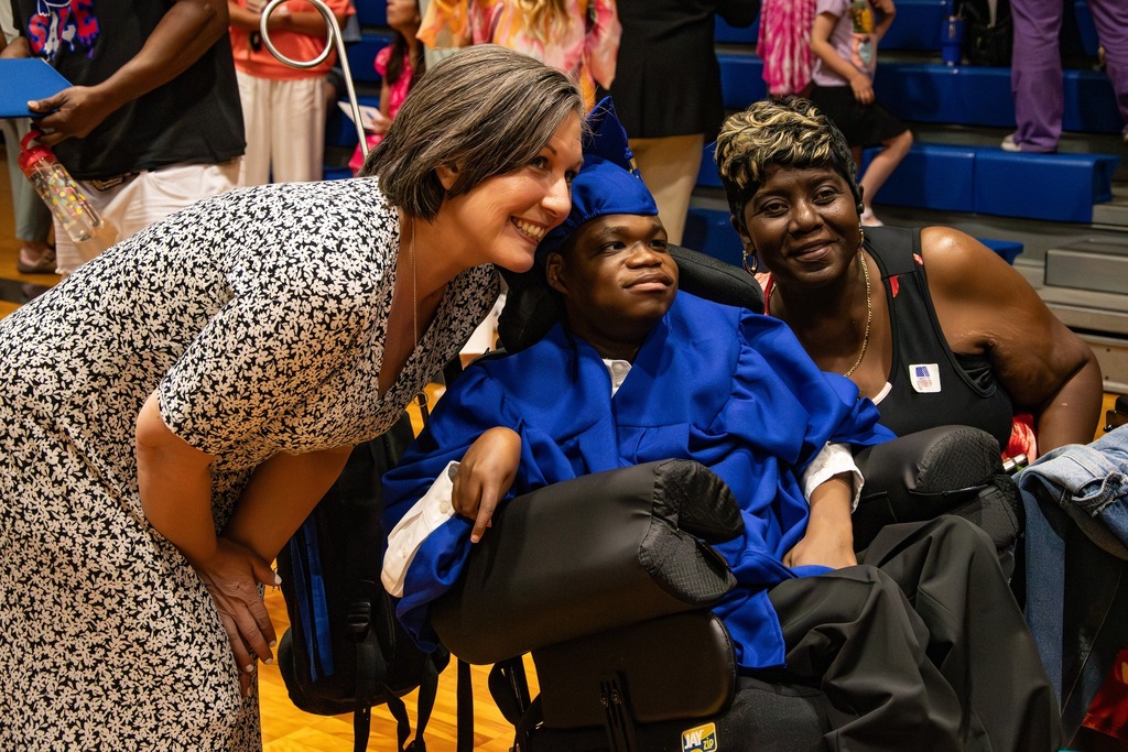 Two women pose for a photo with a Burkett Center graduate following the school's graduation ceremony. The student wears a blue cap and gown.