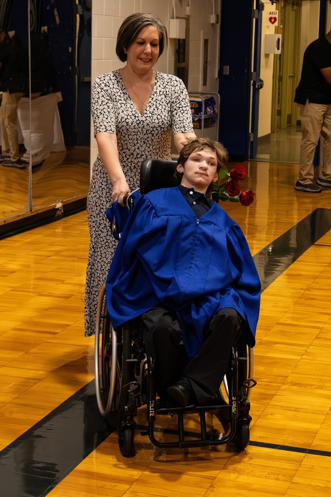 A woman smiles and pushes a Burkett Center graduate in a wheelchair. He wears a blue graduation cap and gown.