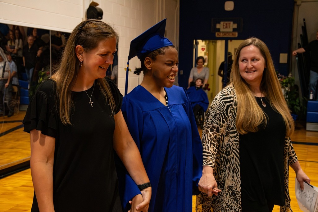 Two women hold the hands of a Burkett Center graduate as she enters the gymnasium for graduation. The student smiles and wears a blue cap and gown.