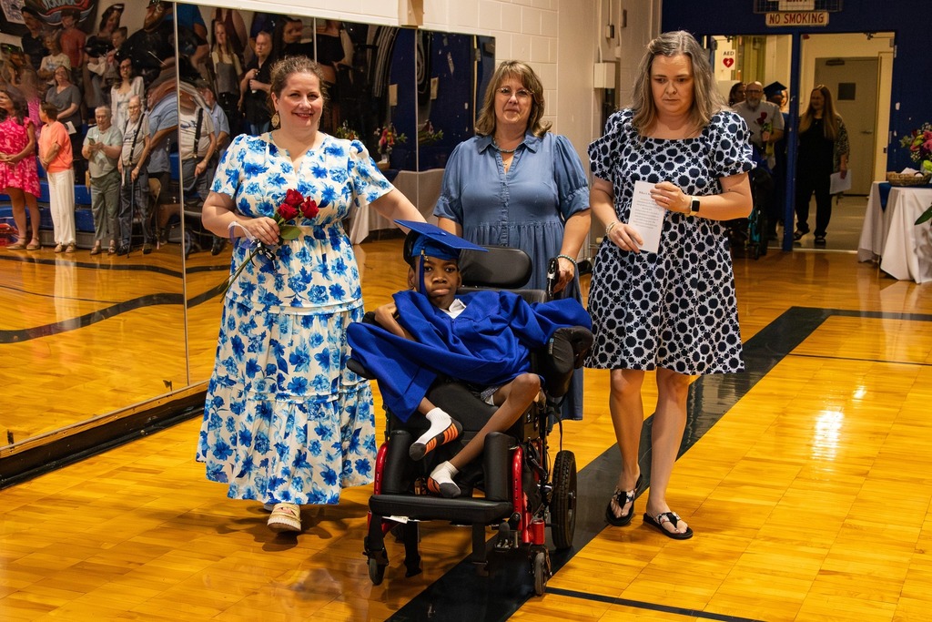 Three women walk behind a Burkett Center graduate as the student enters the gym for graduation. The student wears a blue graduation cap and gown.