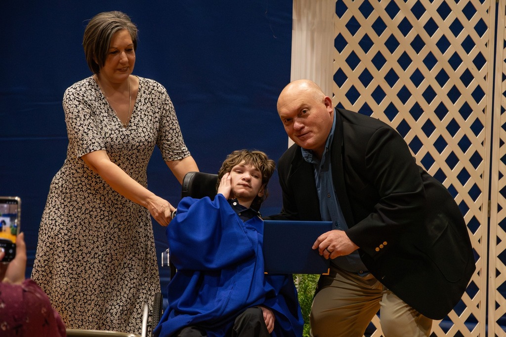 A woman and the Burkett Center principal pose for a photo with a Burkett Center graduate during the school's graduation ceremony. The student wears a blue cap and gown.