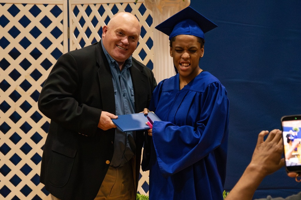 The Burkett Center principal smiles for a photo with a Burkett Center graduate during the school's graduation ceremony. The student wears a blue cap and gown.