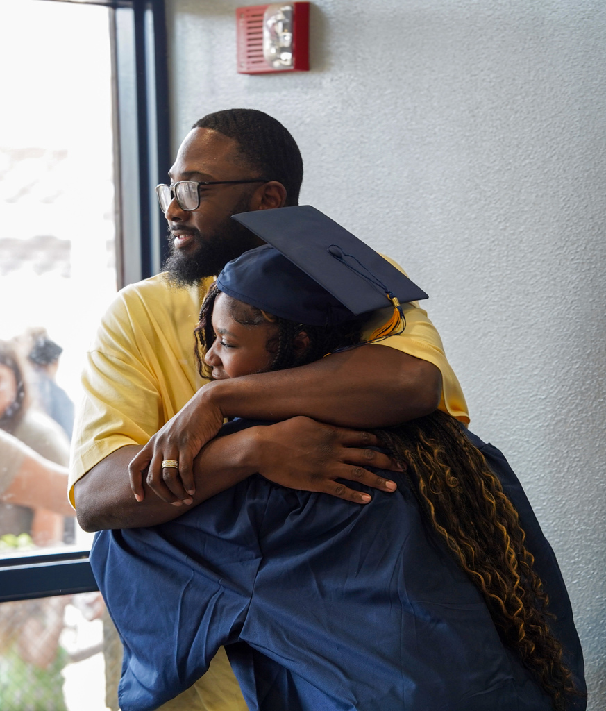 A JCVAL graduate and a man hug following the school's graduation ceremony. The graduate wears a blue cap and gown.