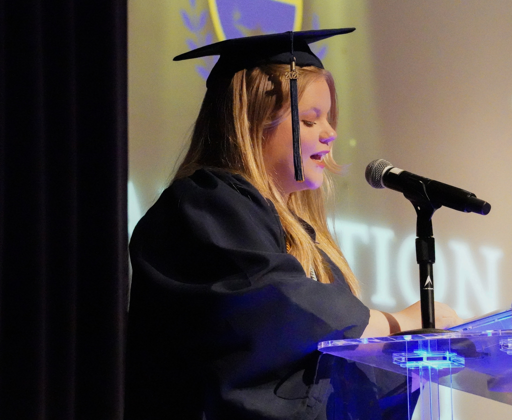 A JCVAL graduate speaks into a microphone during the school's graduation ceremony. The student wears a blue cap and gown.
