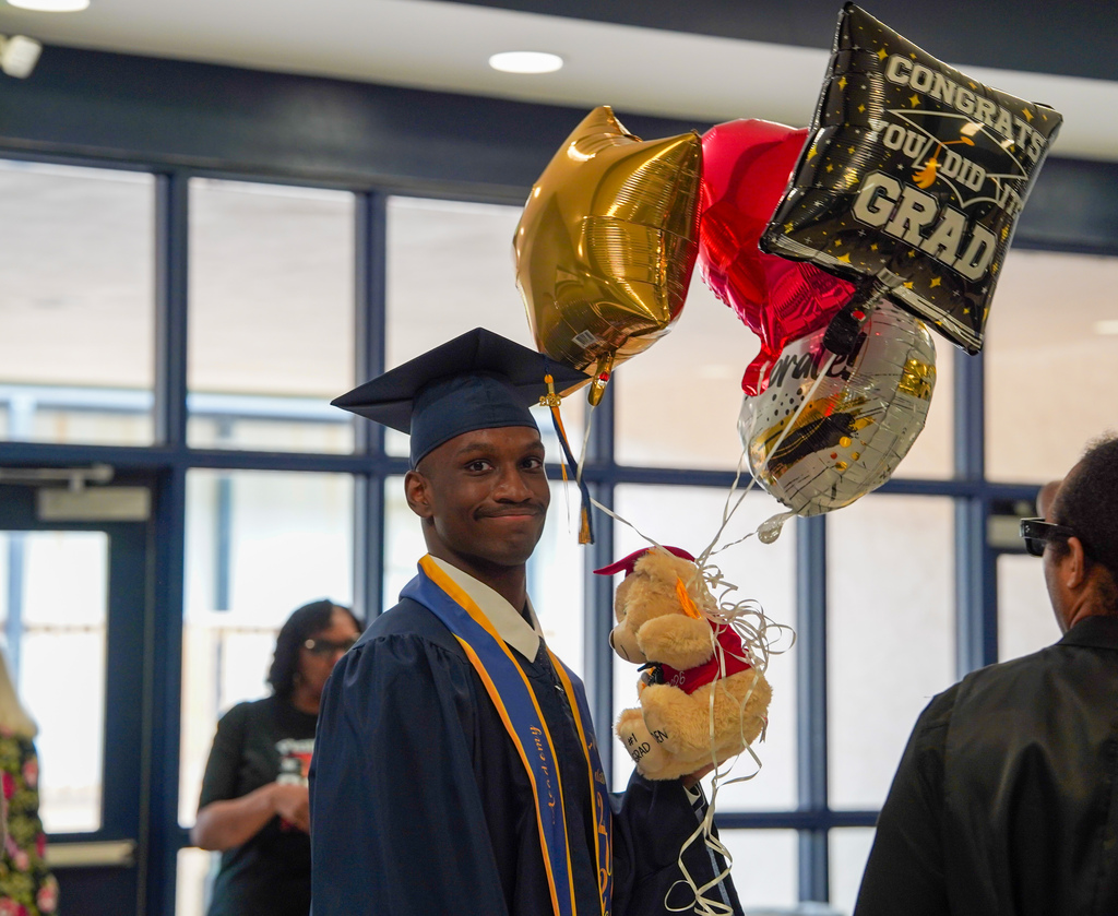 A JCVAL graduate smiles for a photo following the schools graduation ceremony. He holds congratulatory balloons and wears a blue cap and gown.
