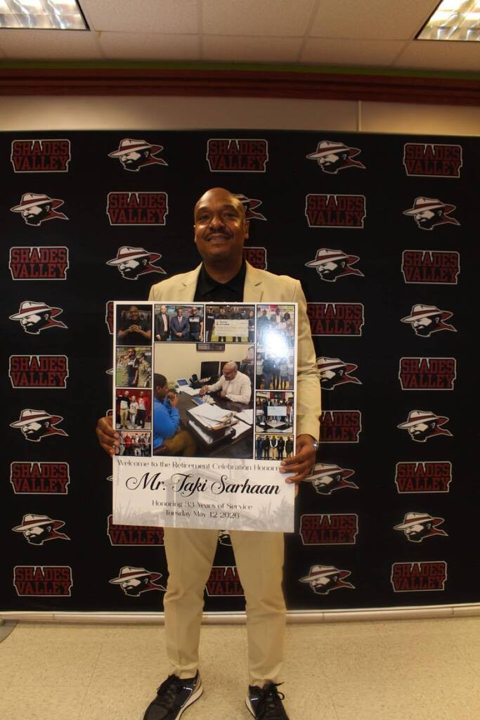 Shades Valley Principal Taki Sarhaan smiles for a photo in front of a Shades Valley photo backdrop. He holds a poster celebrating his retirement.