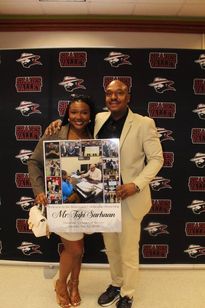 Taki Sarhaan poses for a photo with a woman in front of a Shades Valley High School photo backdrop. They hold a poster celebrating Mr. Sarhaan's retirement.