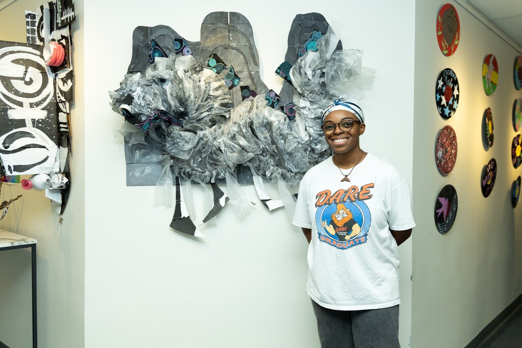 A student smiles for a photo in front of a sculpture made of recycled materials. The sculpture hangs on a wall inside a hallway.