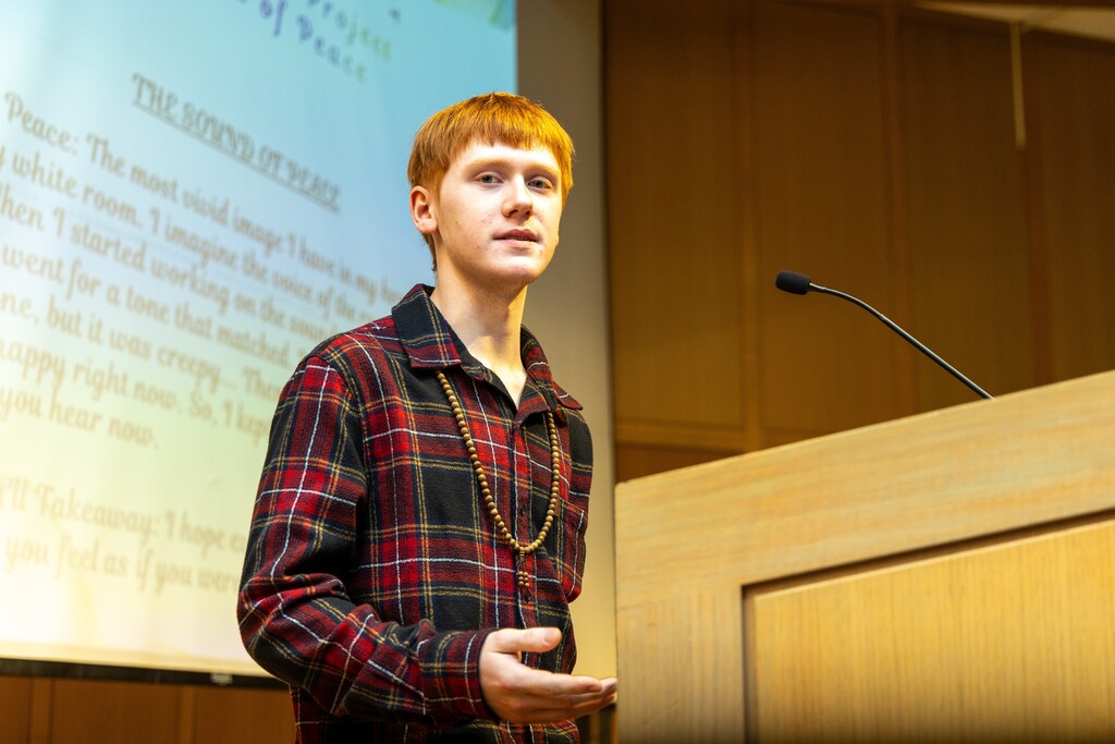 A student speaks into a microphone at a podium while standing in front of a projection on a screen.