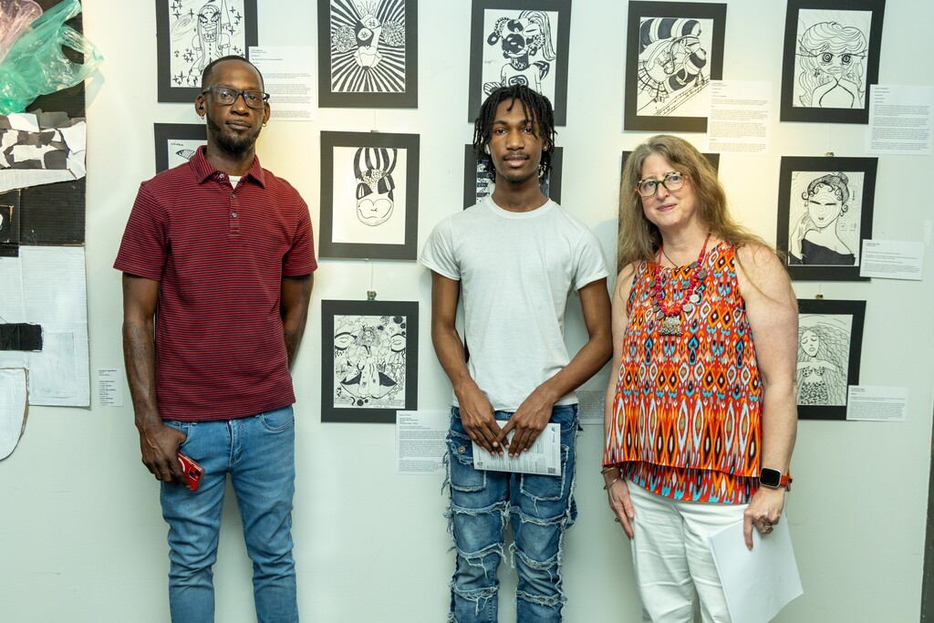 Two adults and a student pose for a photo in front of drawings hanging on a wall at an art museum.