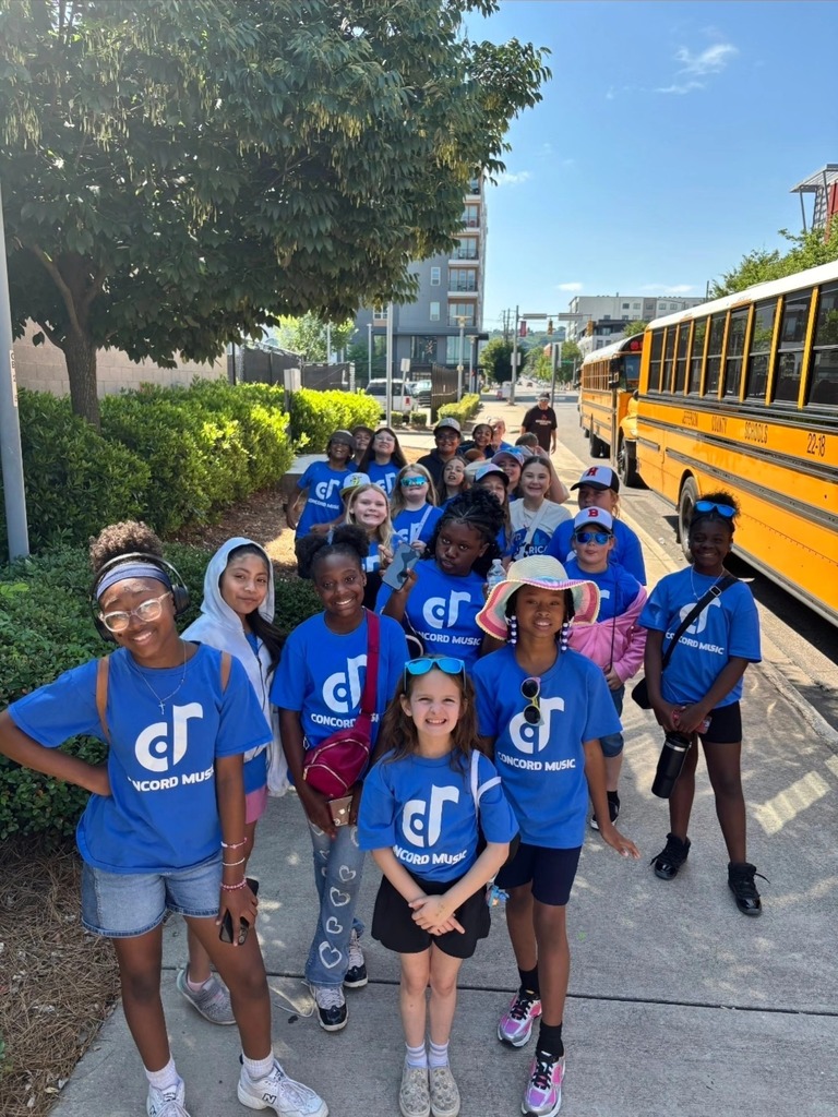 Concord Elementary School choir students stand outside a school bus and smile for a photo. They wear matching blue shirts that say "Concord Music."