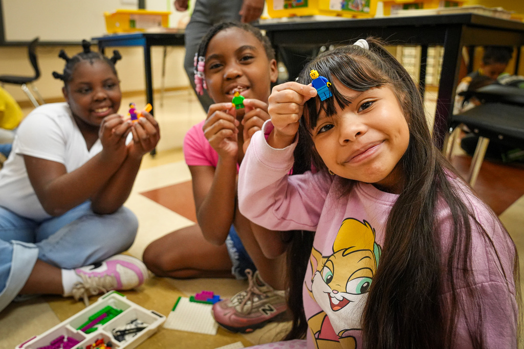 A group of students hold up legos while they sit in the floor of a classroom.