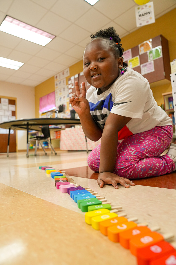 A student holds up a peace sign while sitting on the floor. She sits behind colorful, popsicle-shaped wood pieces with letters printed on them.