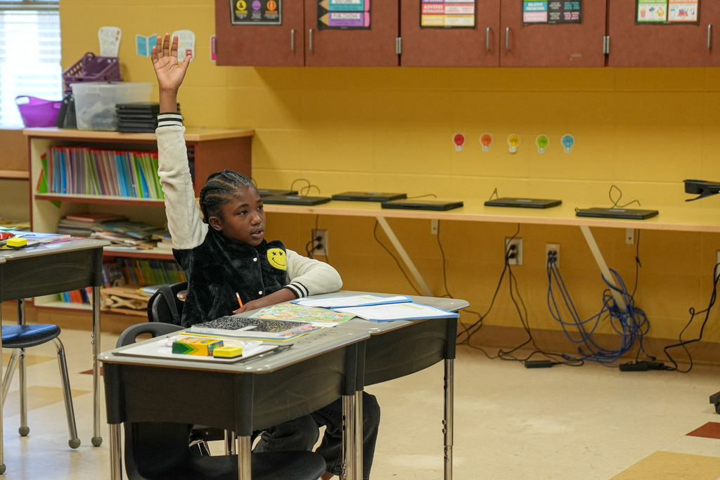 A student raises their hand while sitting at a desk in a classroom.