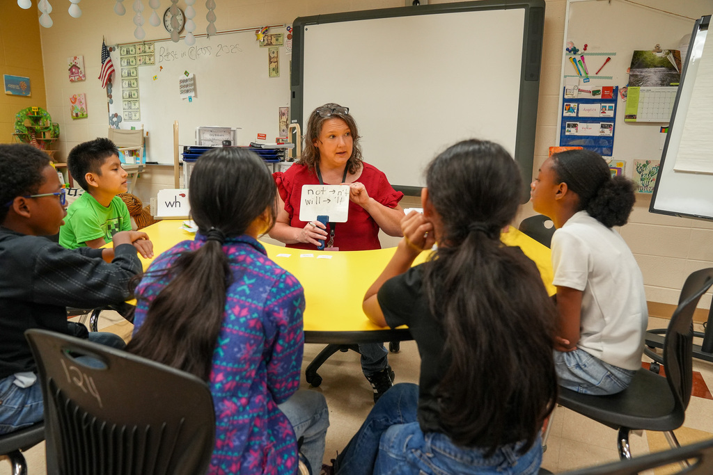A teacher holds up a small white board for students as they sit around her at a table.