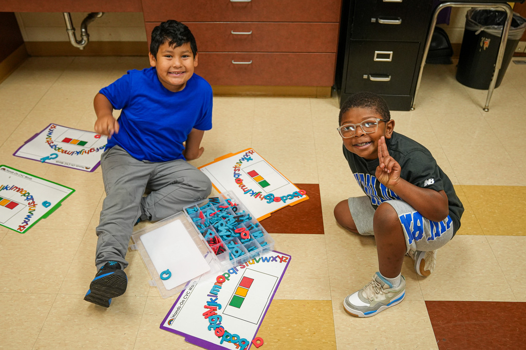 Two boys sit on the floor of a classroom and do an assignment with letter blocks on a mat.