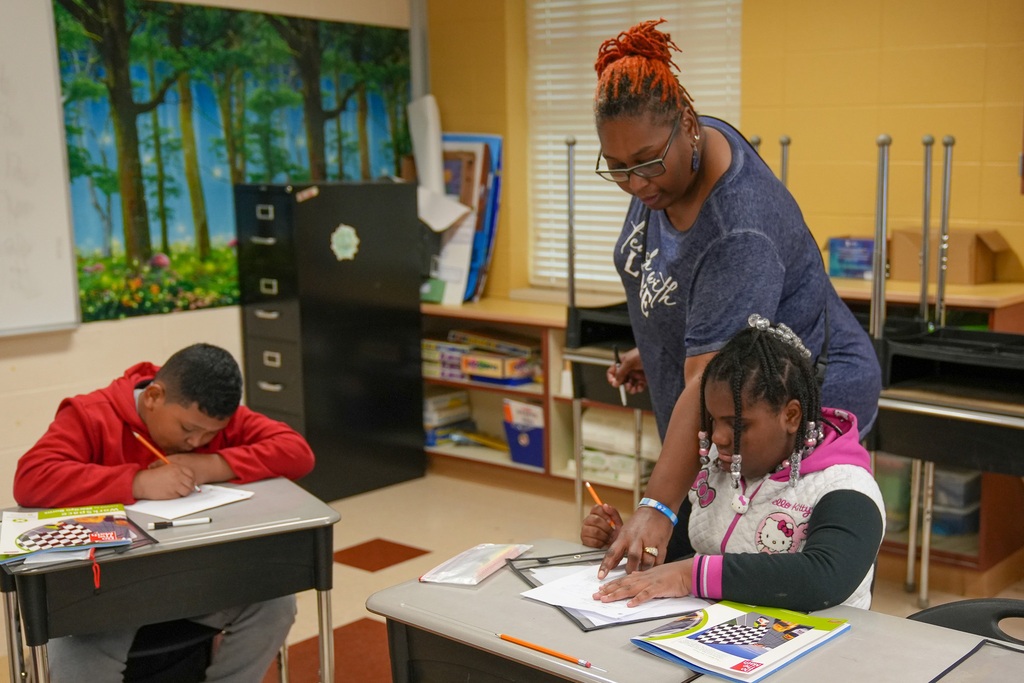 A teacher helps a student with an assignment while the student sits at a desk in a classroom.