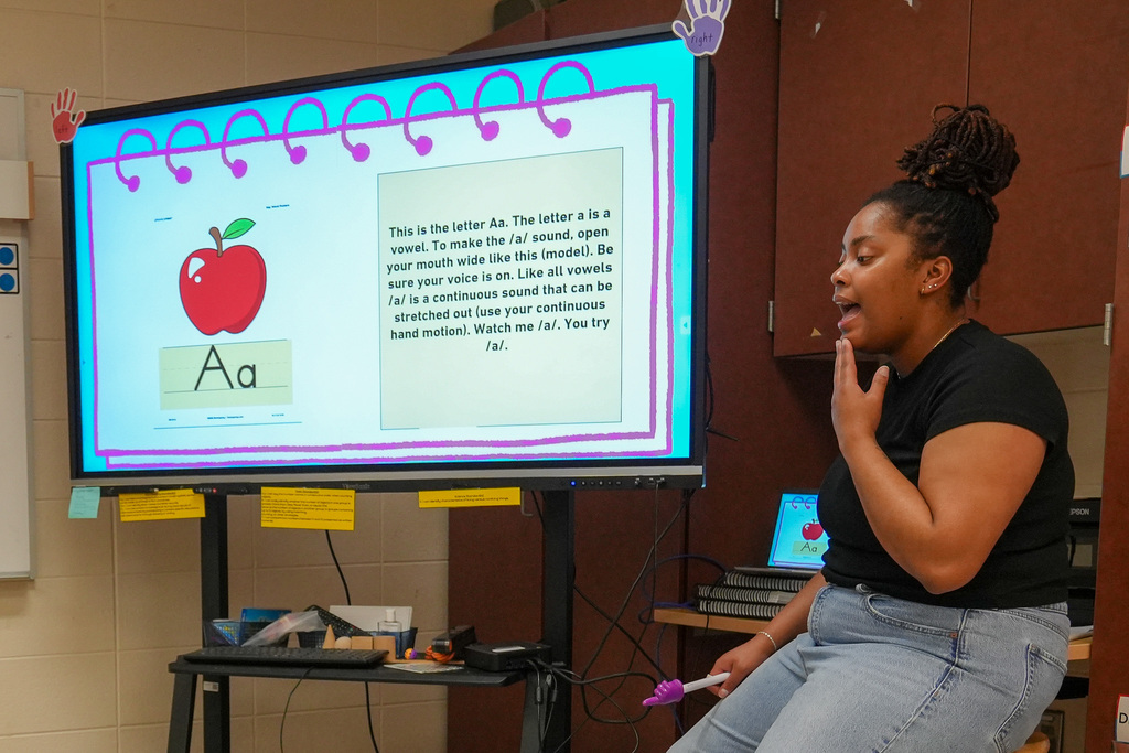 A teacher sits next to a smart board in a classroom and demonstrates how to say the letter 'A'.